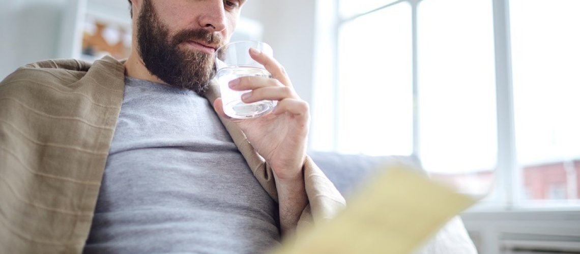 Young man wrapped into blanket drinking water and reading financial paper or letter while staying at home