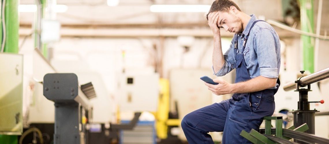 Embarrassed young engineer of factory reading notification in smartphone and touching his forehead