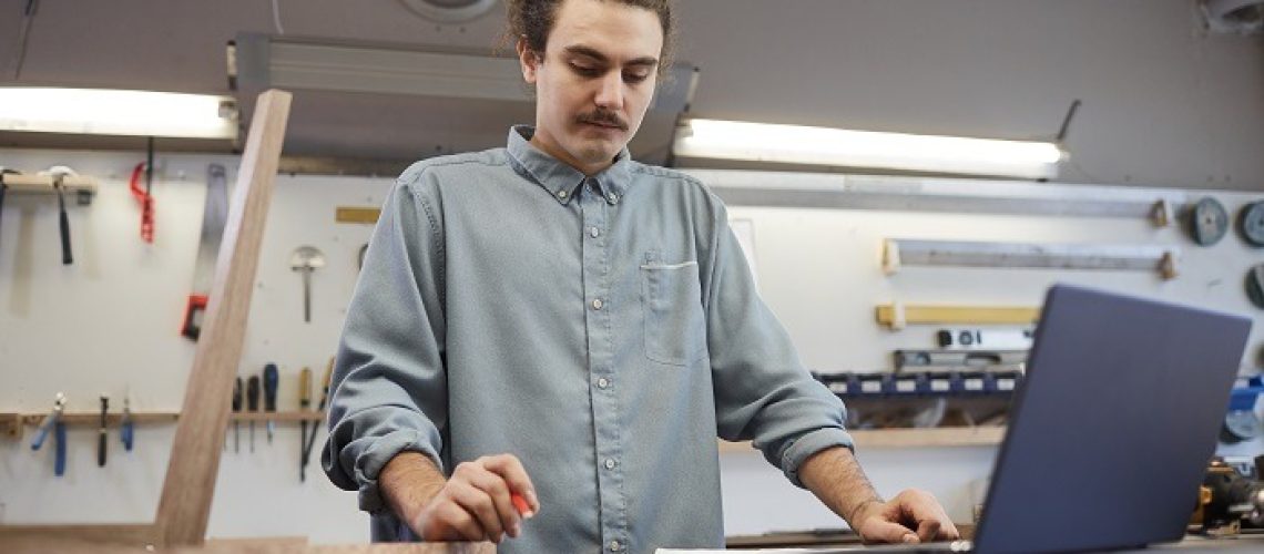 Young businessman standing at the table with laptop and examining document while working in workshop
